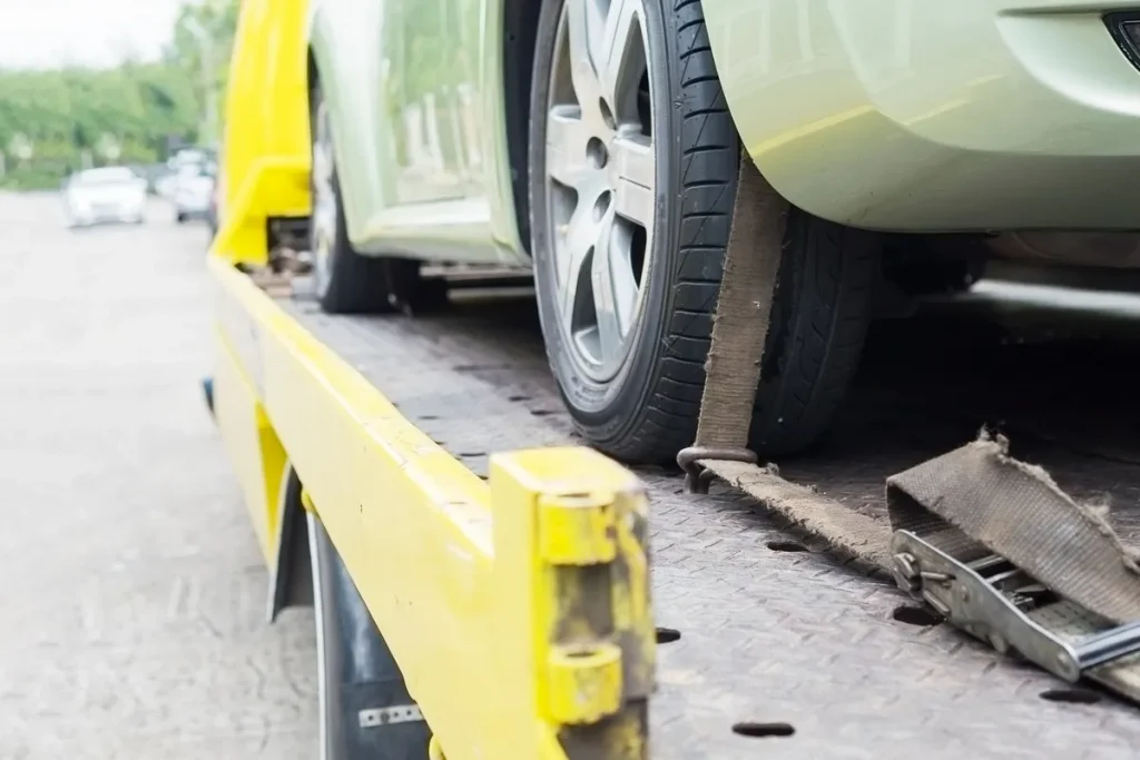 A breakdown lorry transporting a green car with secured belts, illustrating vehicle transport logistics.
