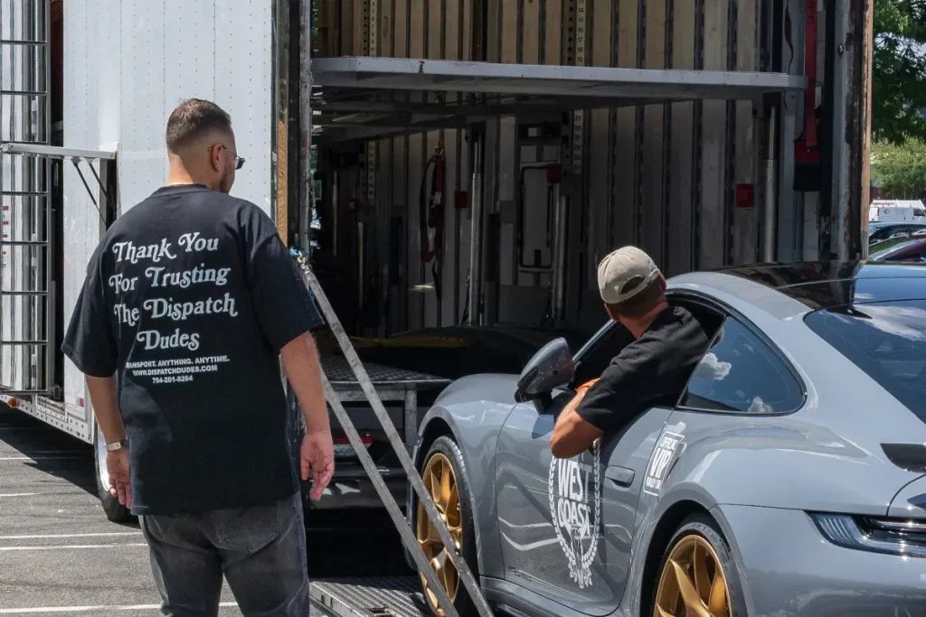 Two men inspecting a gray sports car beside a trailer.